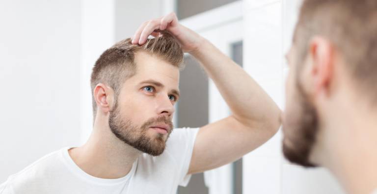 Man with blonde hair and blue eyes wearing white, with attractive facial hair inspecting his hair part for hair loss